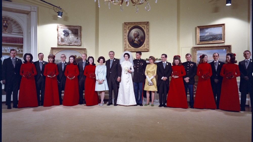 In this photograph by Frank Wolfe, President Lyndon B. Johnson and First Lady Lady Bird Johnson pose with their daughter, Lynda Bird Johnson, on her wedding day on Dec. 9, 1967. The Johnsons are joined by Lynda's groom, Marine Corps Capt. Charles S. Robb, her younger sister, Luci Baines Johnson, to the right of Mrs. Johnson, and the wedding party in the Yellow Oval Room on the second floor of the White House. This photograph was captured following Lynda and Charles' ceremony in the East Room. Along with the bridal ensemble, Geoffrey Beene designed the bridesmaids’ red velvet dresses, which were inspired by Francisco Goya’s 1787-1788 painting "Manuel Osorio Manrique de Zúñiga" and reflected the medieval aesthetic popularized by the 1967 film Camelot.