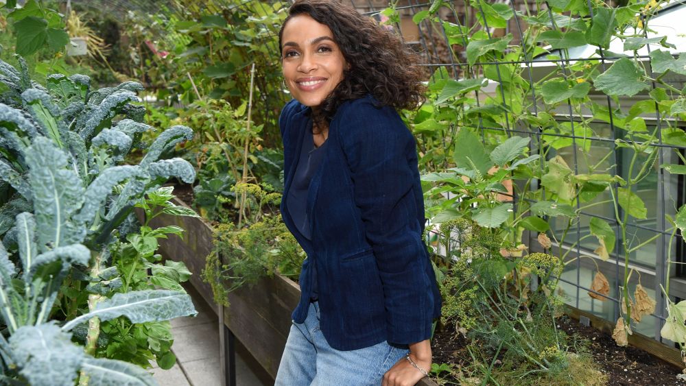 Rosario Dawson in a blue blazer and jeans in a community garden surrounded by lush green vines.